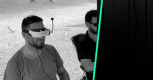 טייס FPV חובש משקף וידאו ומטיס רחפן בשטח פתוח, לצדו נצפה ששומר קשר עין עם הרחפן
